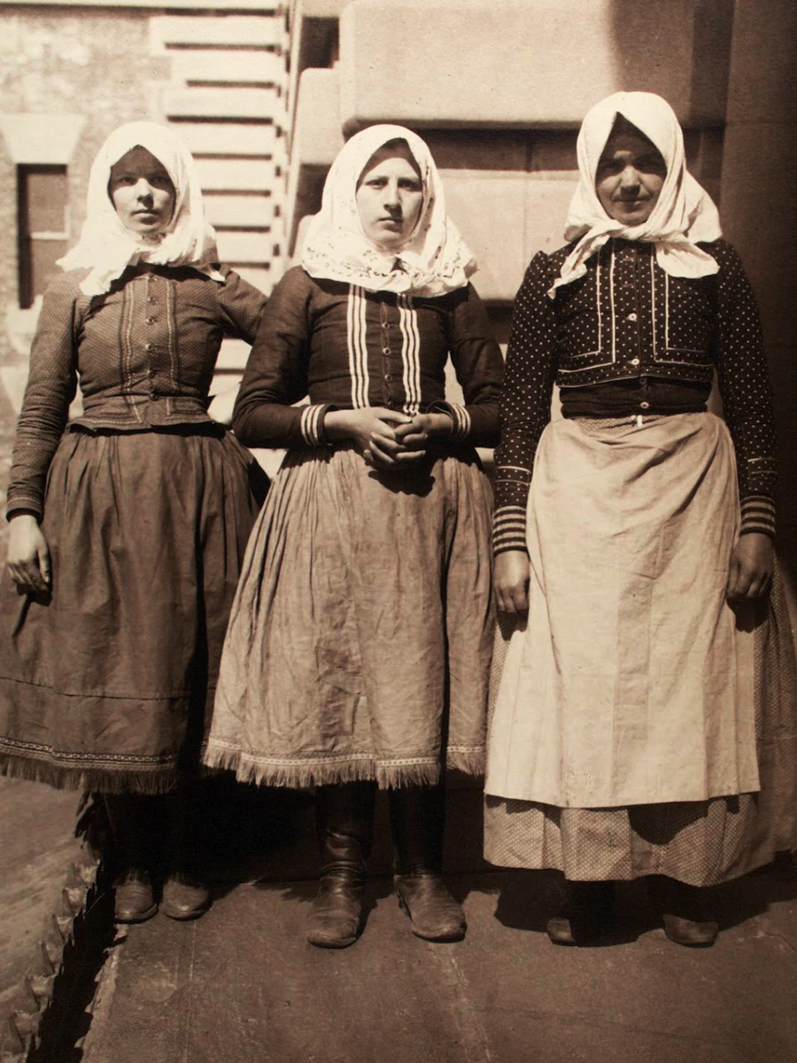 A sepia-toned photo of three young women in traditional clothing, each wearing a headscarf tied under her chin and layered garments, standing together in front of a stepped stone structure.