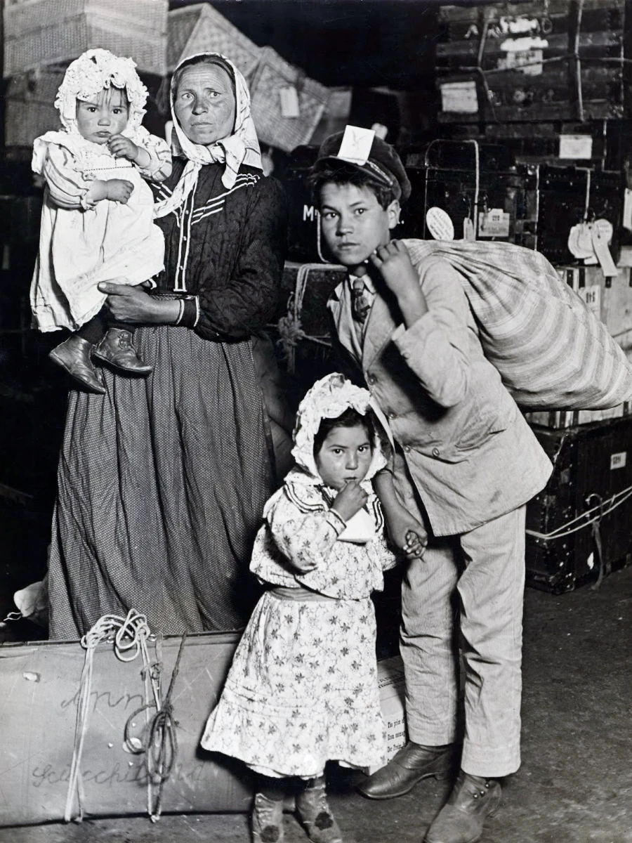 A black and white photo of a family of four amidst a backdrop of luggage and crates. A mother holds her baby with a serious expression and dark clothes, beside her stands a young boy with short hair, wearing a cap, and a young girl sucking on candy, wearing a floral dress and a bonnet.