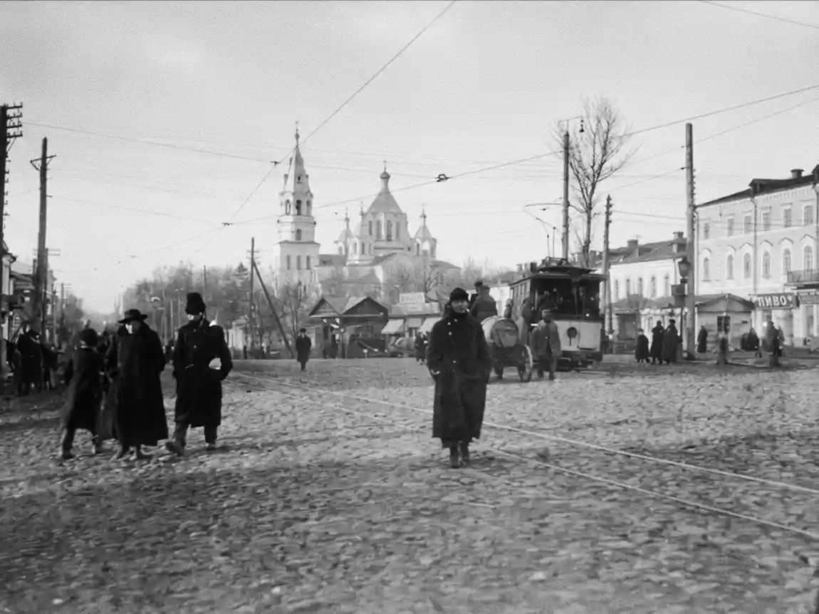 A black and white photo of a street in Dnipropetrovsk, Russia. Pedestrians walk in long coats, while a tram and a church are set along a cobblestone road in the background.