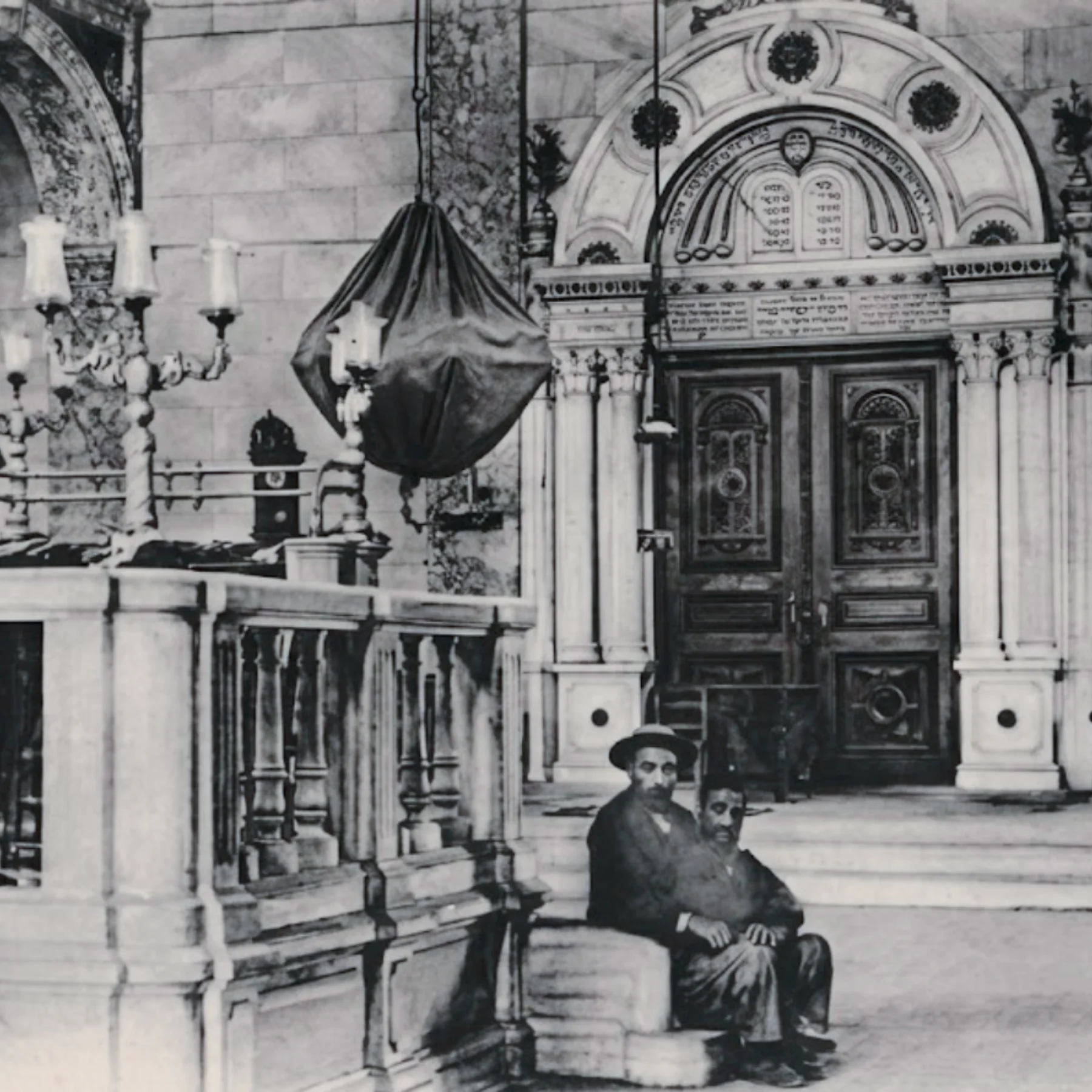 Black and white photo of two men sitting on a bench inside an ornate building with decorative columns and an arched doorway in the background.