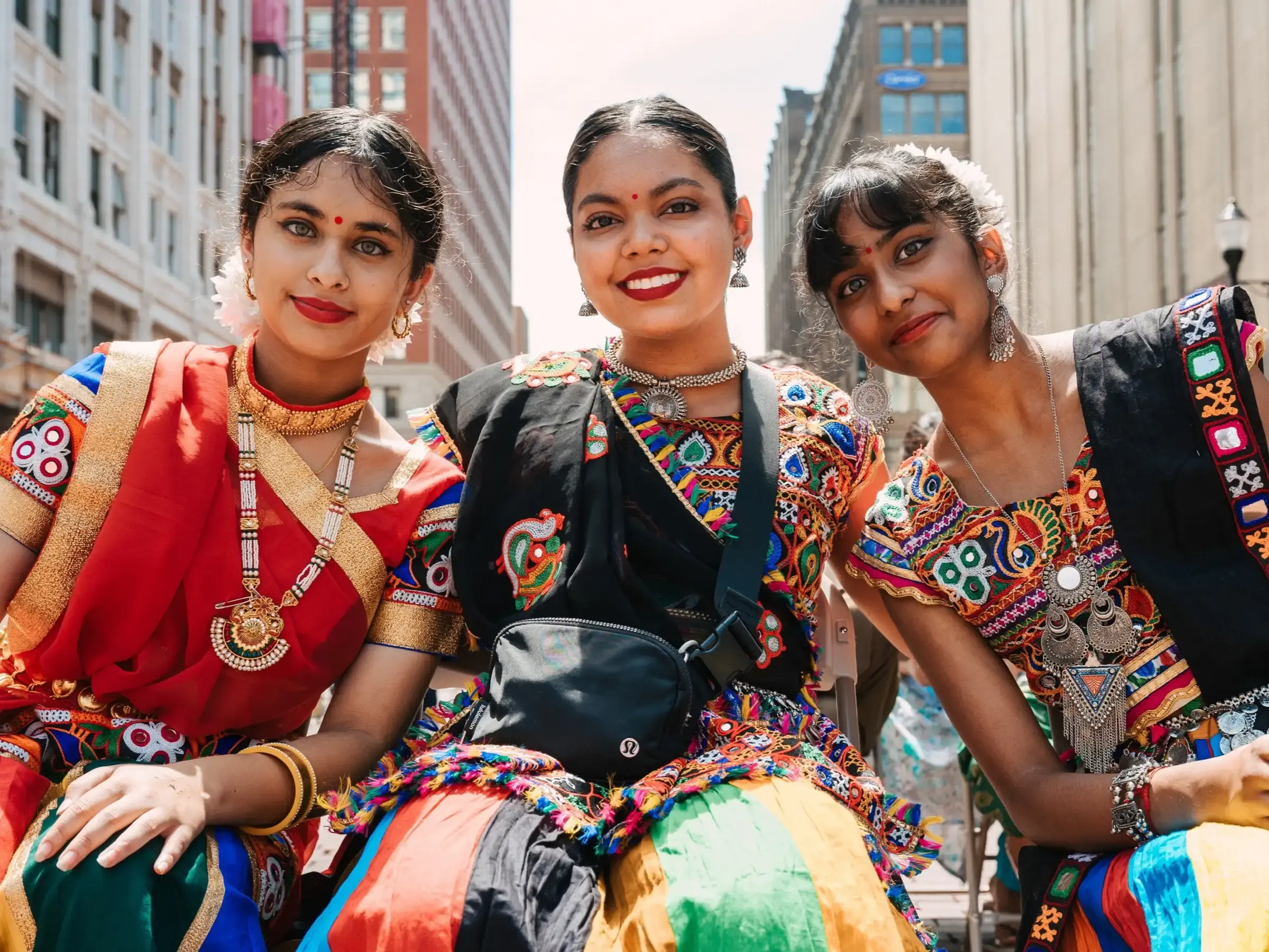A close-up photo of three young, tan-skinned women in colorful traditional attire as they celebrate India's Independence Day, smiling joyfully against an urban backdrop.