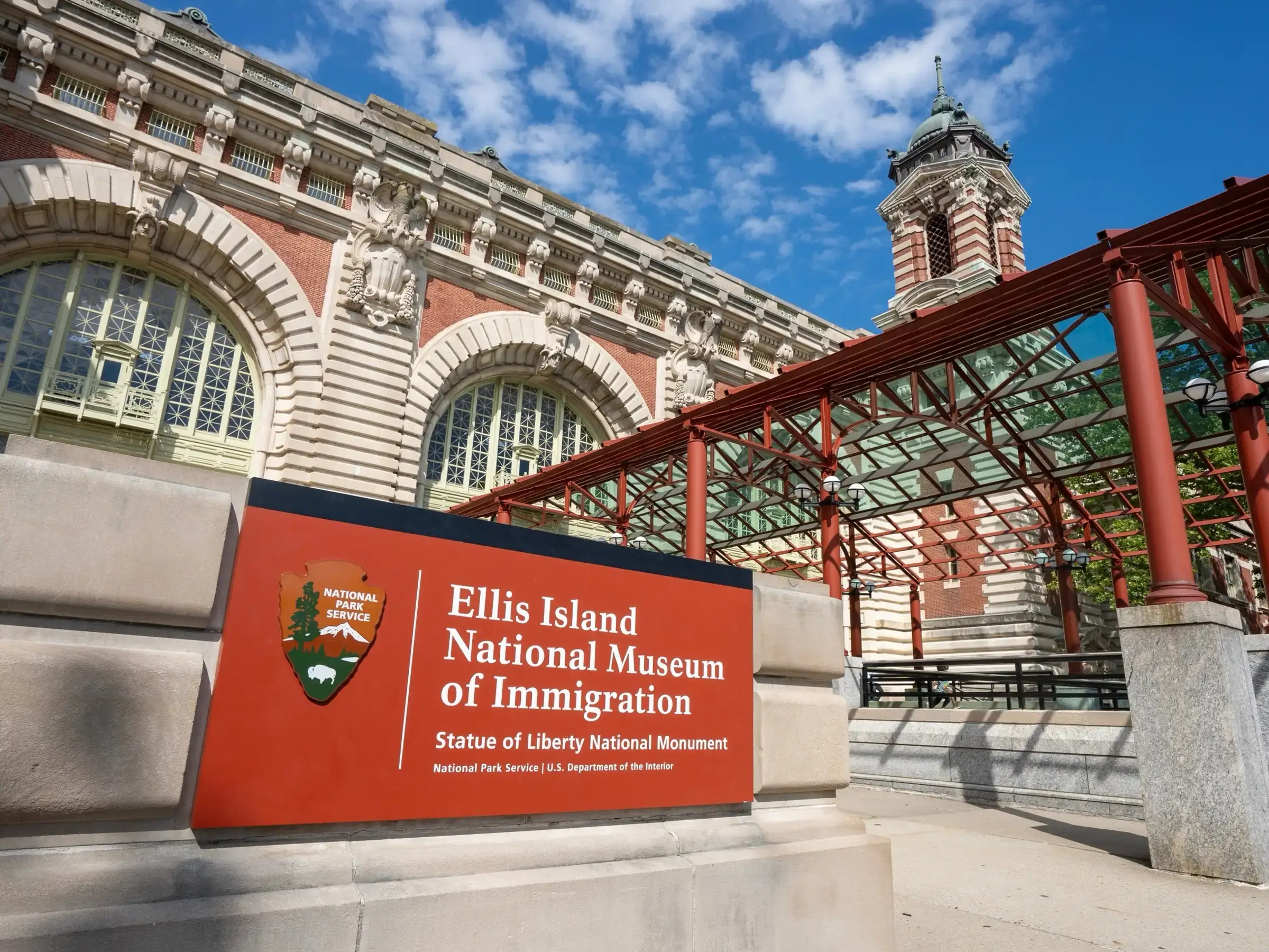 The exterior of the Ellis Island National Museum of Immigration with a red brick facade, large arched windows, and a clock tower against a blue sky. A red sign on the floor in front hosts the museum’s name and National Park Service emblem.