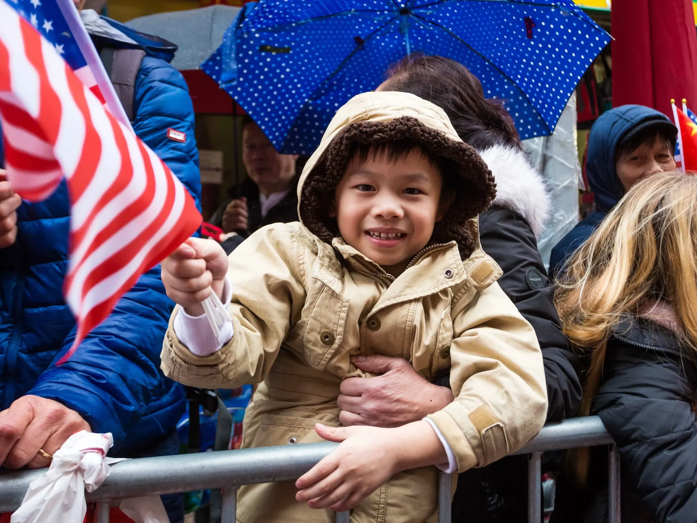 A light-skinned, smiling child in a tan coat waves an American flag at a modern NYC Chinese Lunar New Year parade, surrounded by adults and colorful umbrellas.