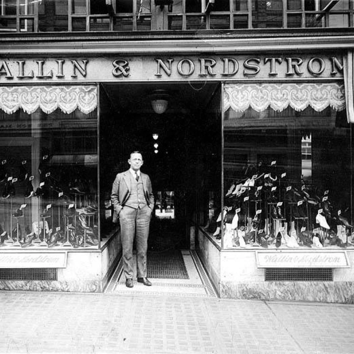 Black and white early 20th century photo of a man in a suit standing in front of the "Ballin & Nordstrom" shoe store. Various shoes are displayed through large glass windows.