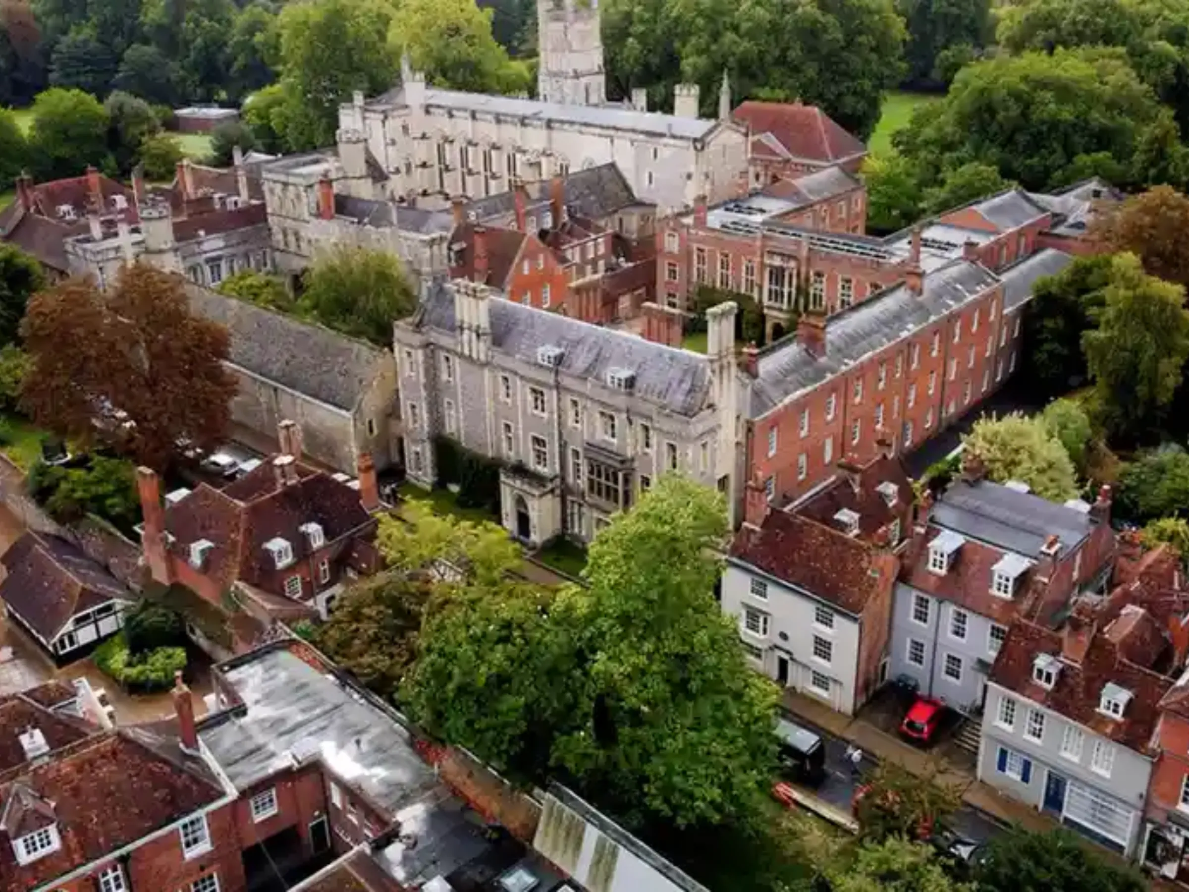 Aerial view of Winchester College’s building complex on a sunny day with stone and red brick structures. Green trees are both around and throughout the campus.
