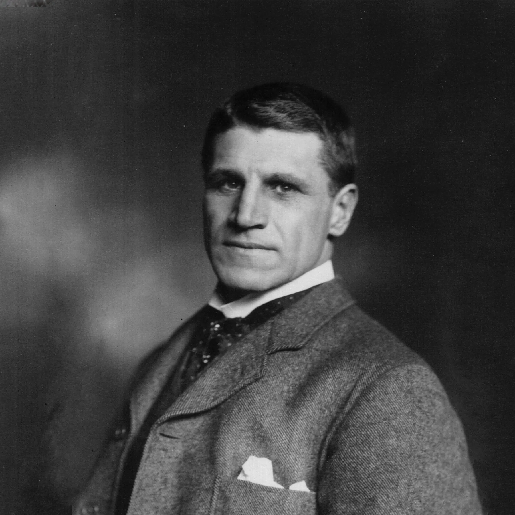 Black and white portrait from the late 1800s of a light-skinned man. His body is turned slightly to the left while his face is turned toward the camera. He wears a suit jacket with a handkerchief in the breast pocket, a white dress shirt, and a cravat.