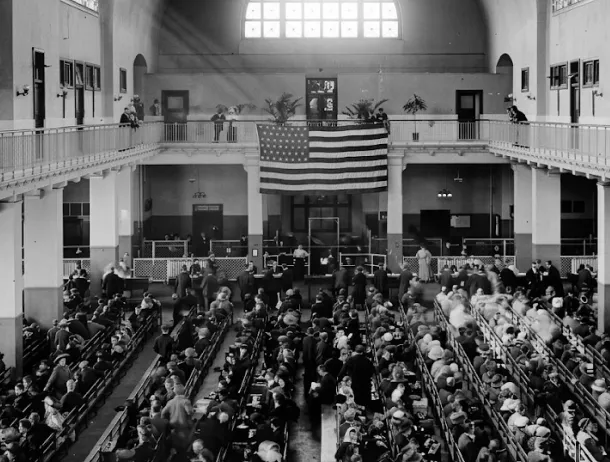 A black and white 20th-century photo overlooks the interior hall of the Ellis Island Main building filled with rows of seated immigrants, and an American flag hanging in the center.