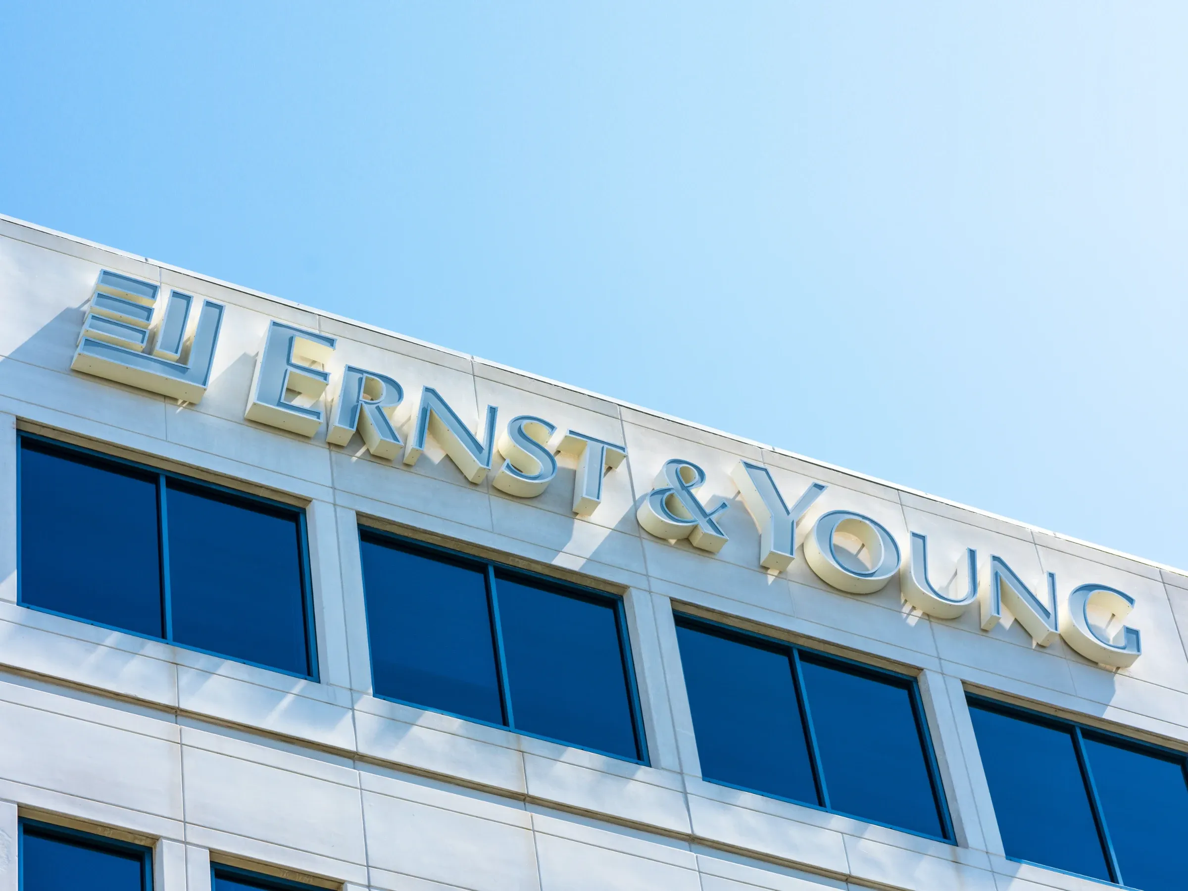 Color photo angled upward at a modern office building with a white exterior featuring the corporate signage for Ernst & Young. Beneath the sign are four windows, and blue sky occupies the top half of the image.