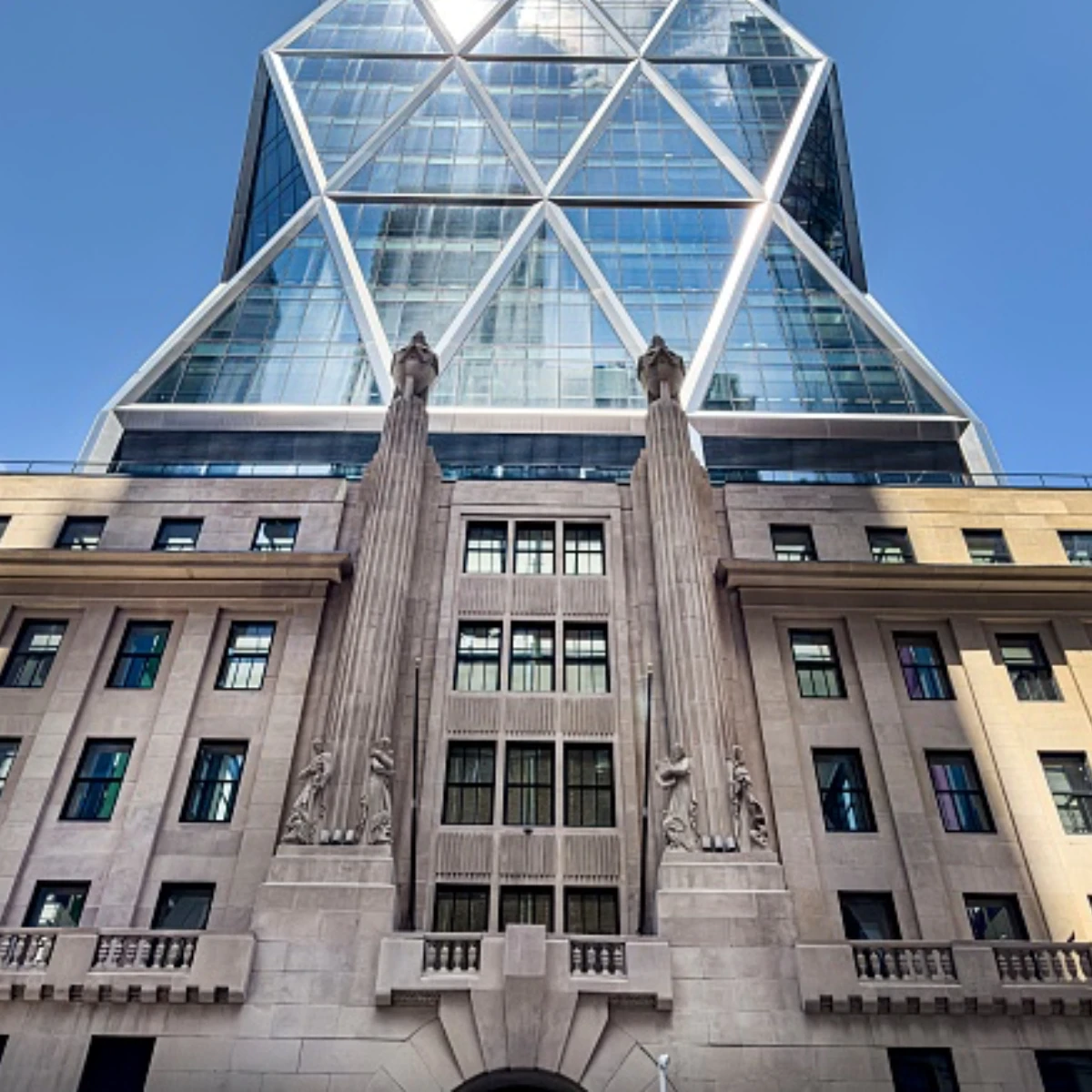 The beige stone facade of Hearst Tower with art deco sculptures beneath a modern glass skyscraper. It has a prominent, repeating triangular steel frame pattern backed by a bright blue sky.