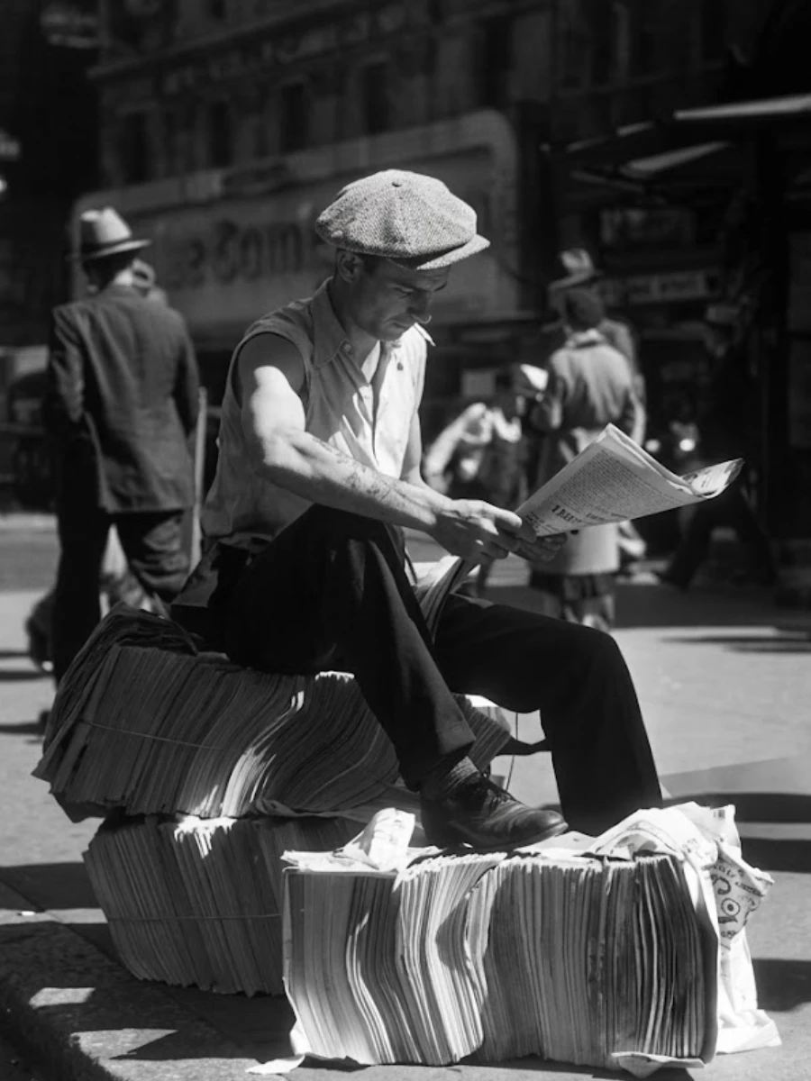 Black and white photo of a man in a flat cap and collared shirt sitting on bundles of newspapers on a city street. He reads one as pedestrians walk in the background.