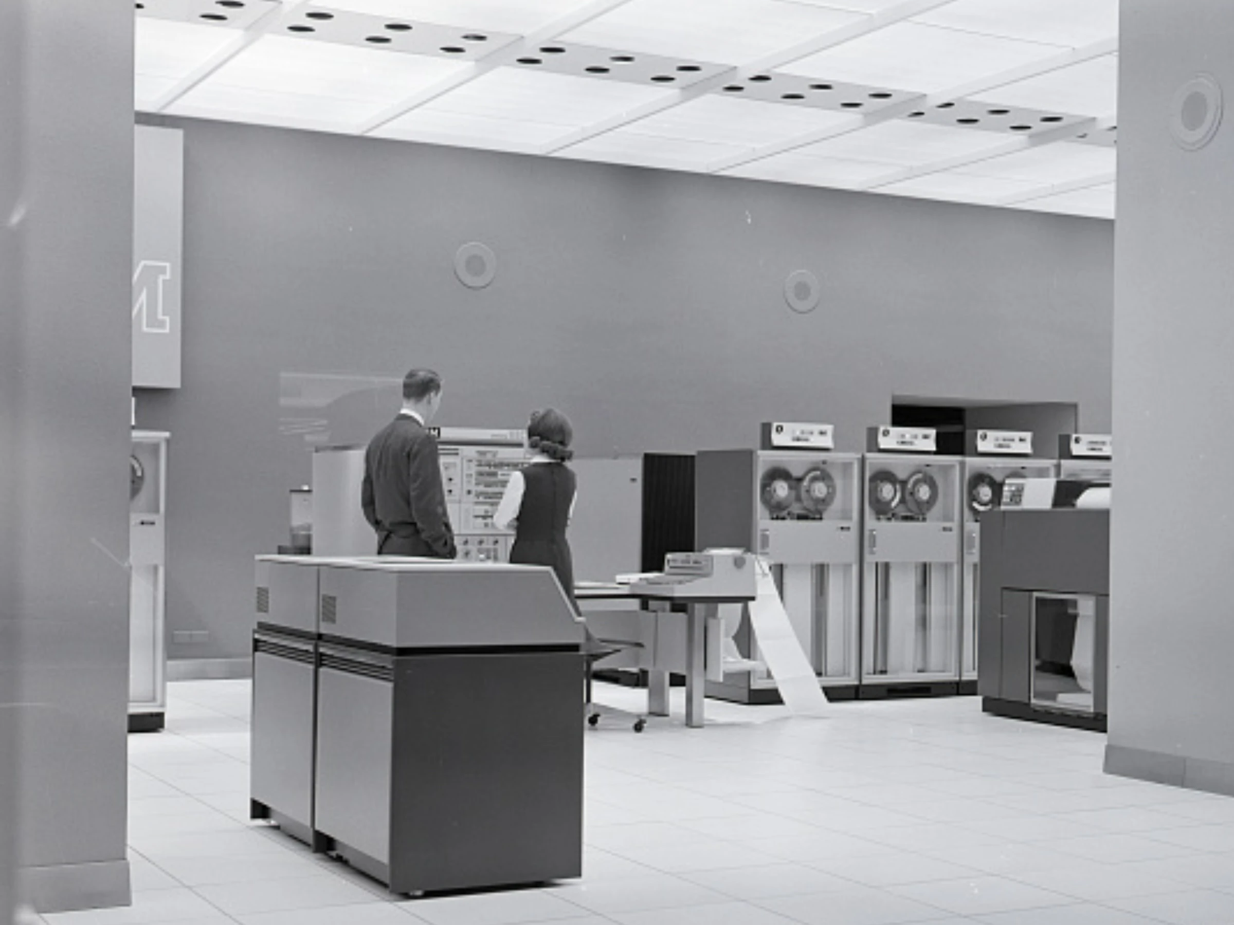 Black and white photo of a mid-20th-century computing facility. In the distance of a dull, gray room, two people stand near a computer printer, with other large machines in the background.