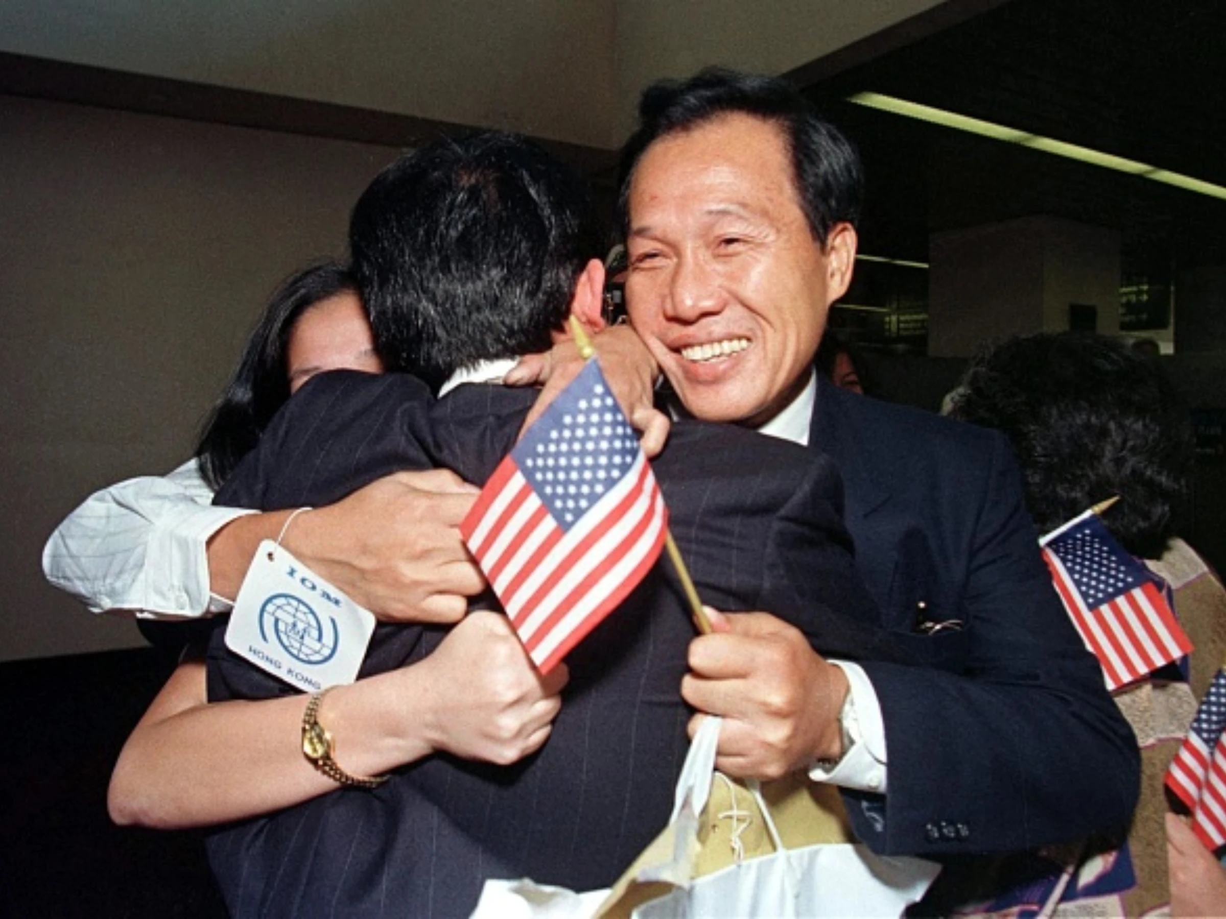 A smiling, light-tan-skinned man in a dark suit and a person whose face is obscured hug a third person in a black suit jacket whose back is facing the camera. The smiling man holds a small United States flag.