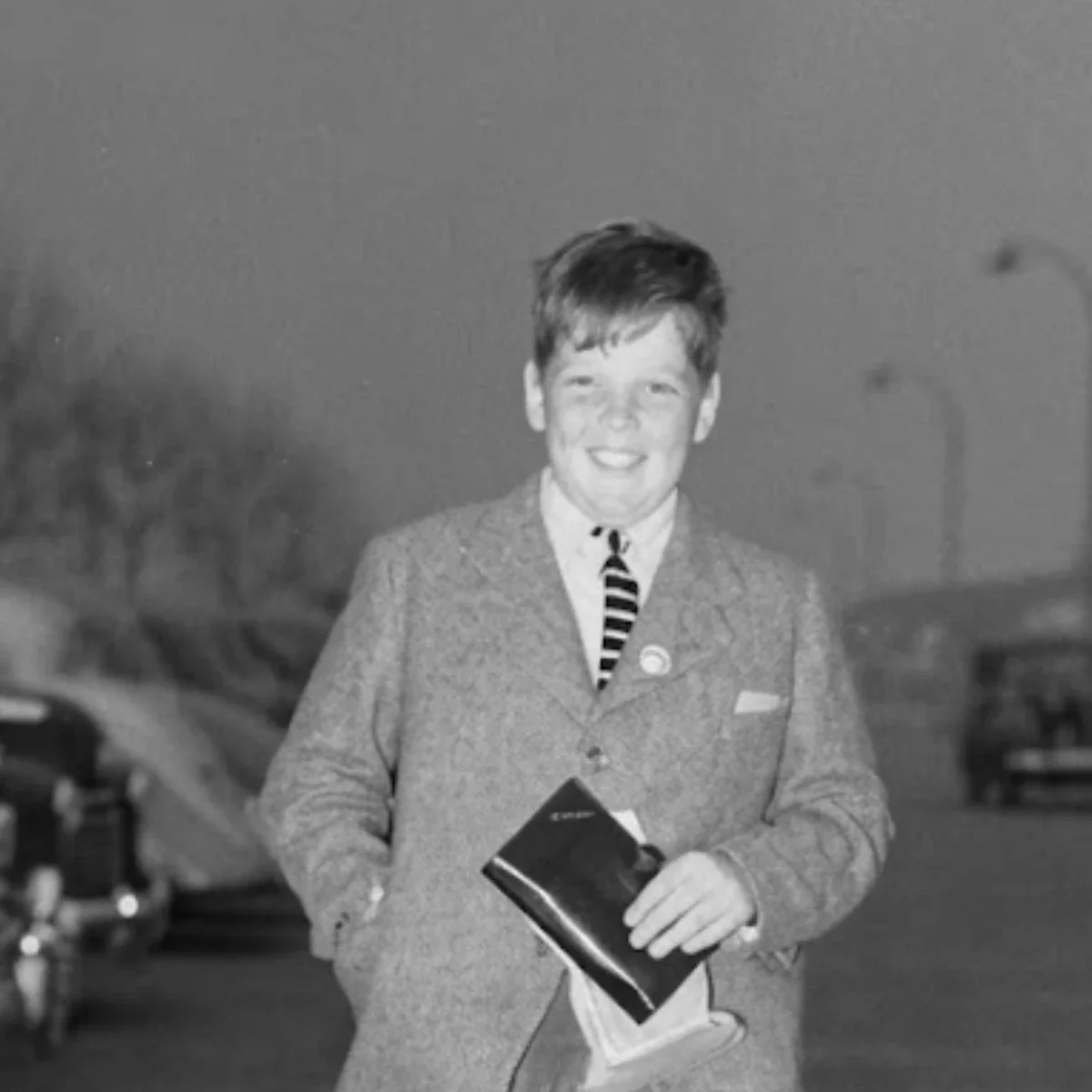 A young, light-skinned Michael Donovan smiling in a black-and-white photograph. He wears a school uniform and holds a dark notebook or booklet. Cars are parked behind him.