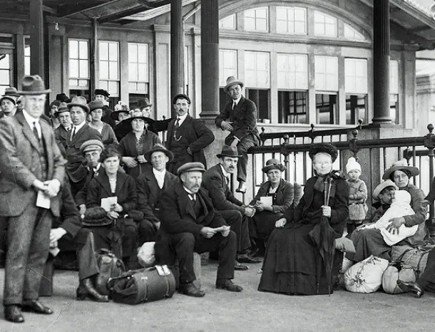 A black and white group photo of early 20th-century light-skinned immigrants of various ages outside a building with their belongings. Some are standing and others are seated. One man sits on the railing.