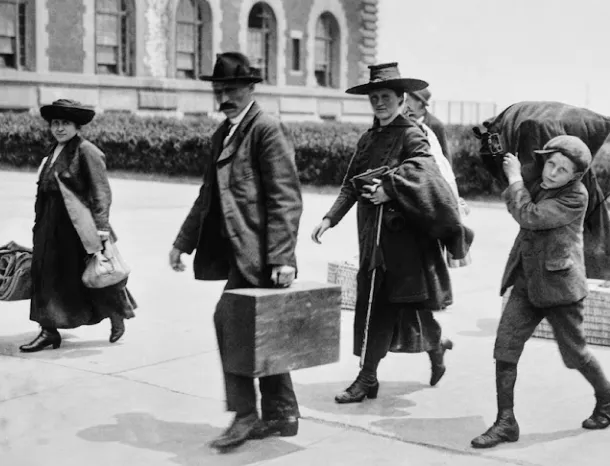 A black and white 20th-century side-facing photo of four immigrants walking with luggage outside a large brick Ellis Island building.