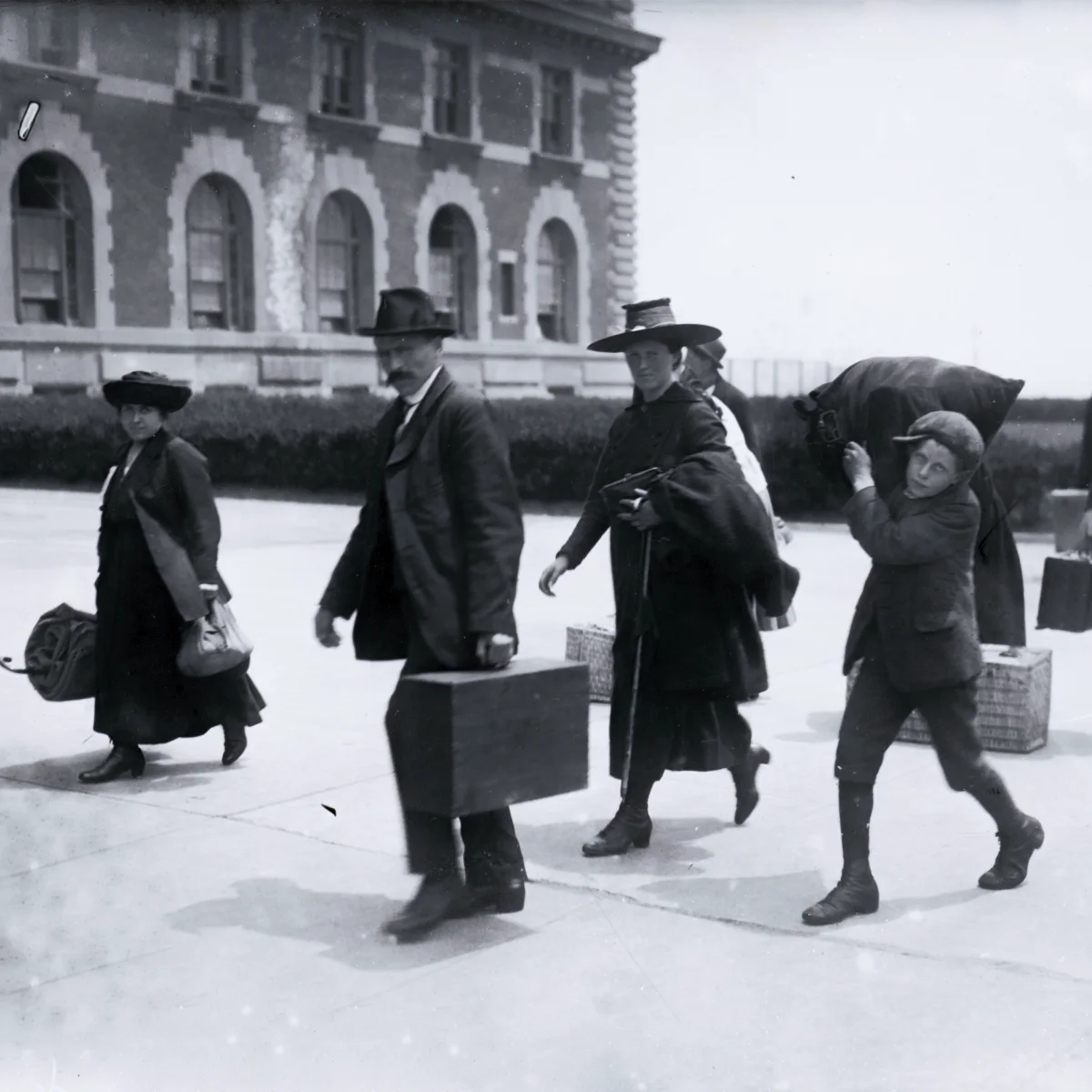 A black and white side-facing photo of four early 20th-century immigrants walking with luggage outside a large brick Ellis Island building.