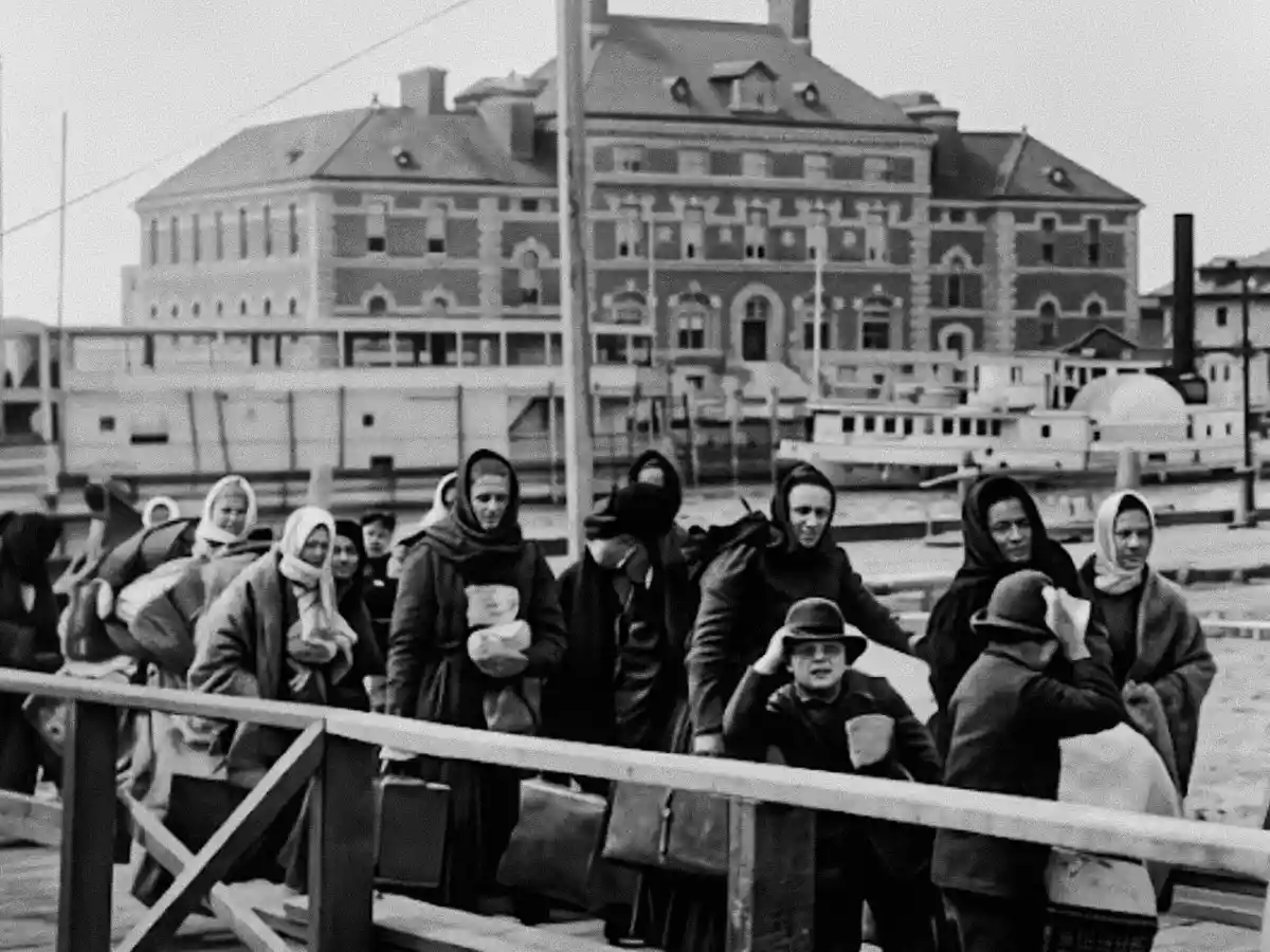 A black and white photo of a group of immigrants, primarily women in heavy coats and headscarves, arriving at Ellis Island and carrying their belongings. A large brick building and boats are visible in the background.