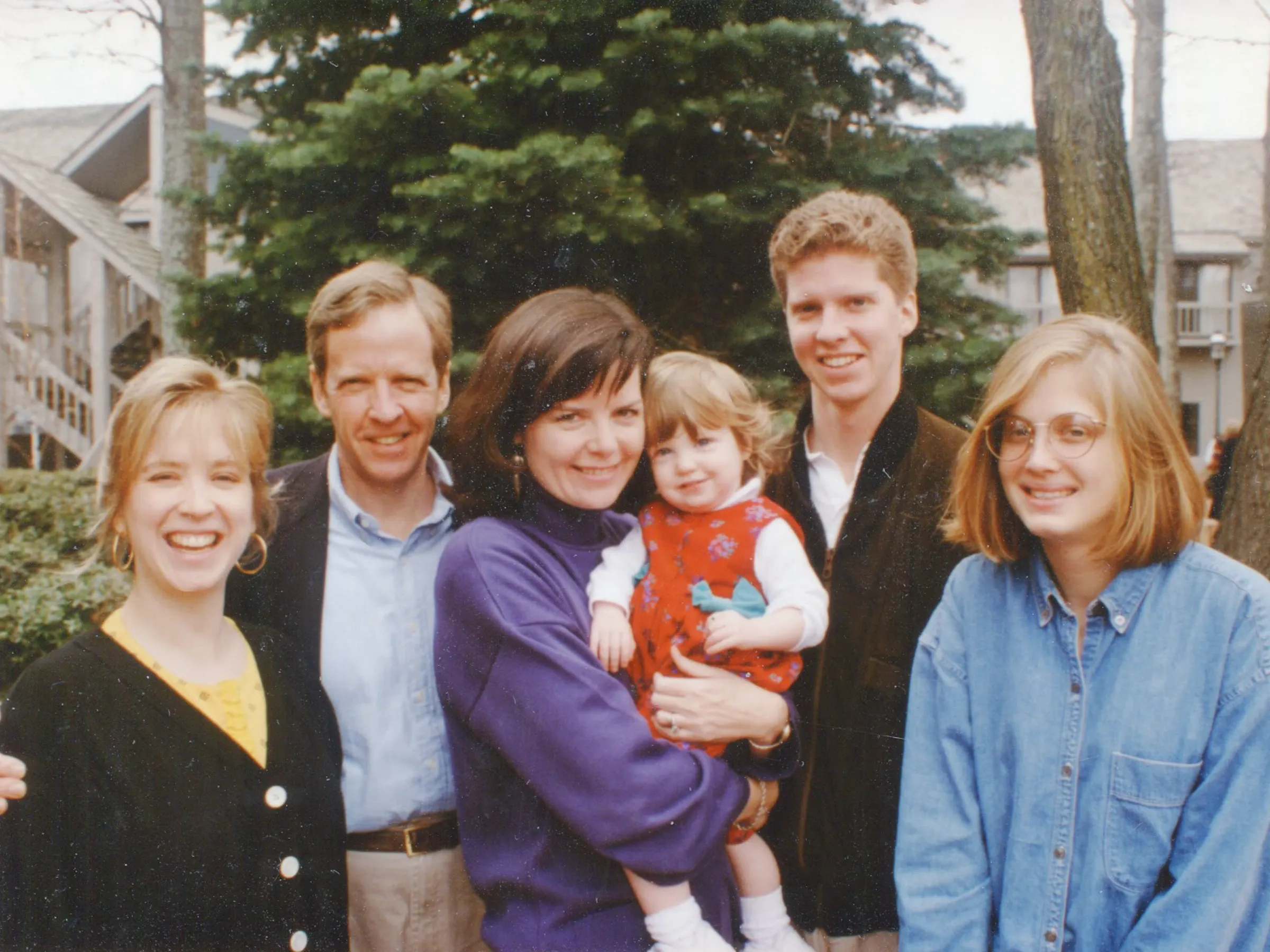 Color photo taken on a sunny day outdoors of Michael and Linda Donovan with four members of their family, all light-skinned. Linda, centered, holds a toddler in a red outfit.