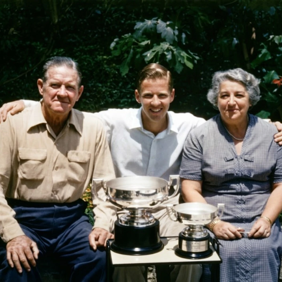 Mid-20th century color photo of Michael Donovan smiling seated on a sunny day outdoors between his mother on the right and his father on the left, all light-skinned. His hands are outstretched on their shoulders. There are trees behind them and two silver trophies in front of them on a small table.