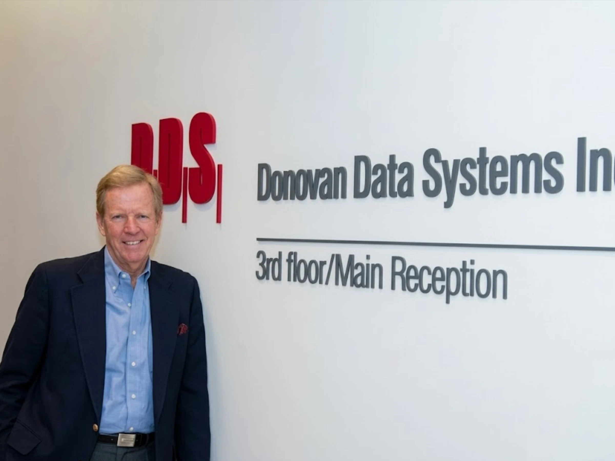 Image of Michael Donovan, a middle-aged white man, wearing a suit jacket and light blue shirt, leaned against a white wall with Donovan Data Systems logo on it.