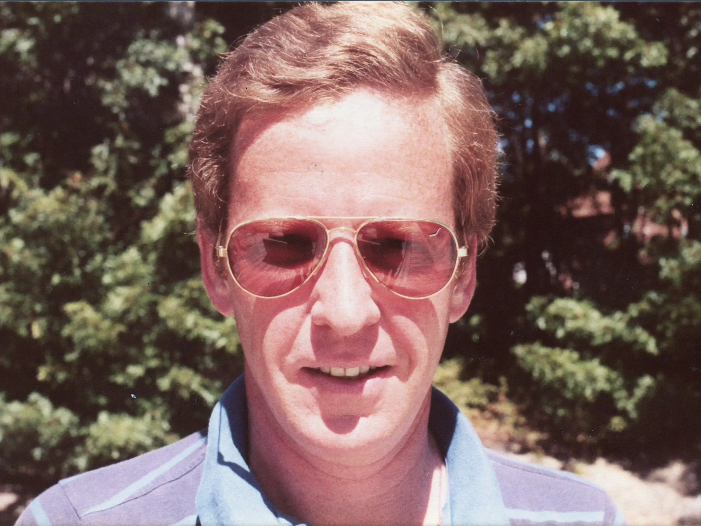 Close up photo of a white man with red hair, wearing Aviator-style sunglasses with red-tinted lenses and a shirt with a blue collar. There are dense green plants behind him.