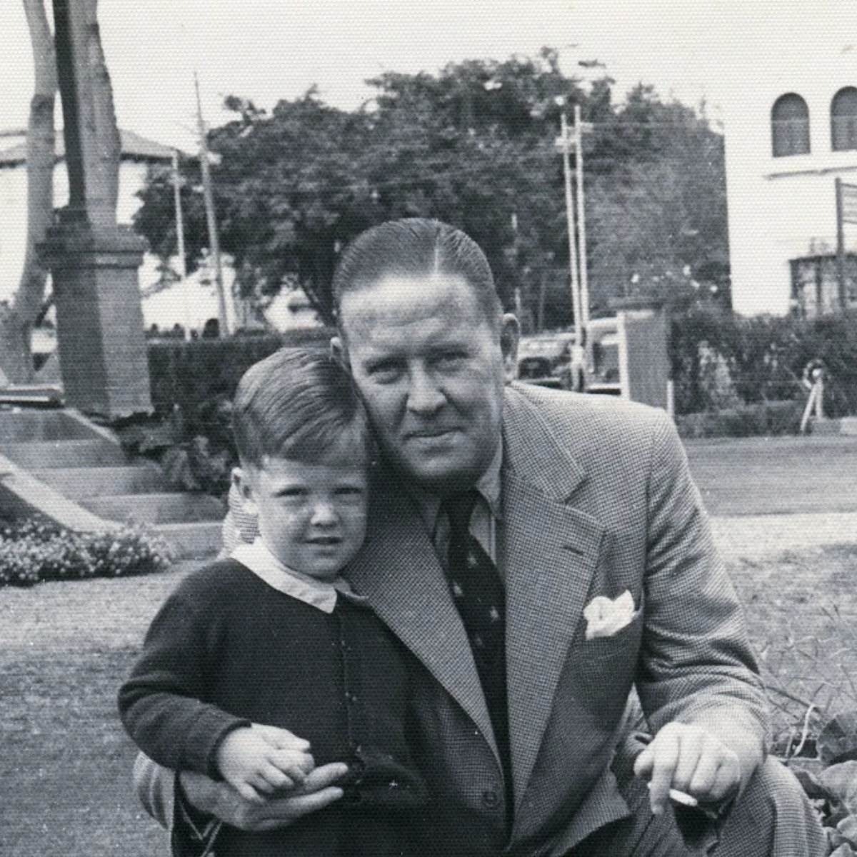 Black and white photo, of a young, light-skinned boy and adult man outdoors. The boy stands wearing a dark sweater. The man, in a suit, crouches and hugs the boy with one arm, holding a cigarette in the other.