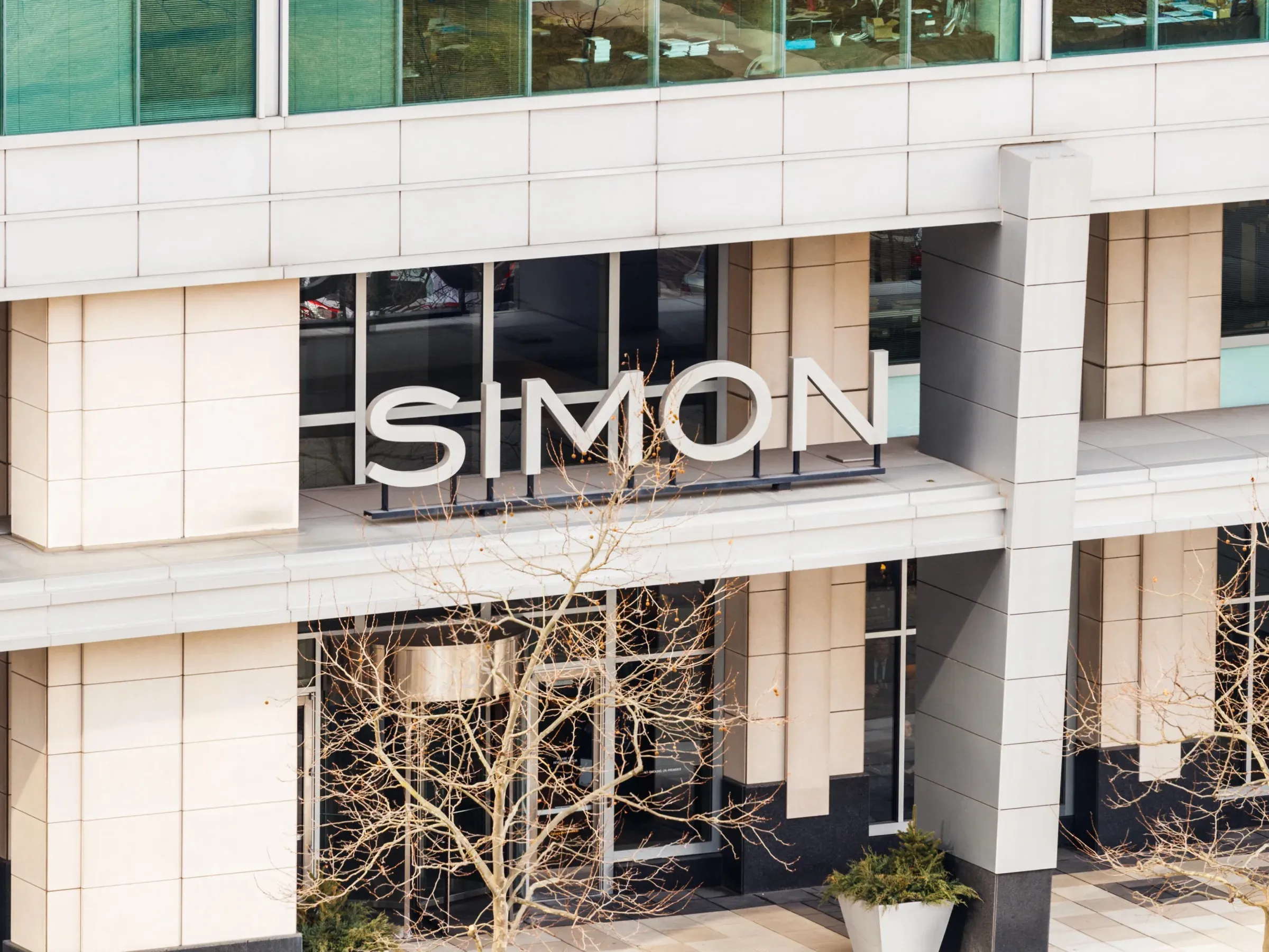 Color photo of a modern building entrance featuring the name "SIMON" in large white letters above glass doors and below green-tinted windows.