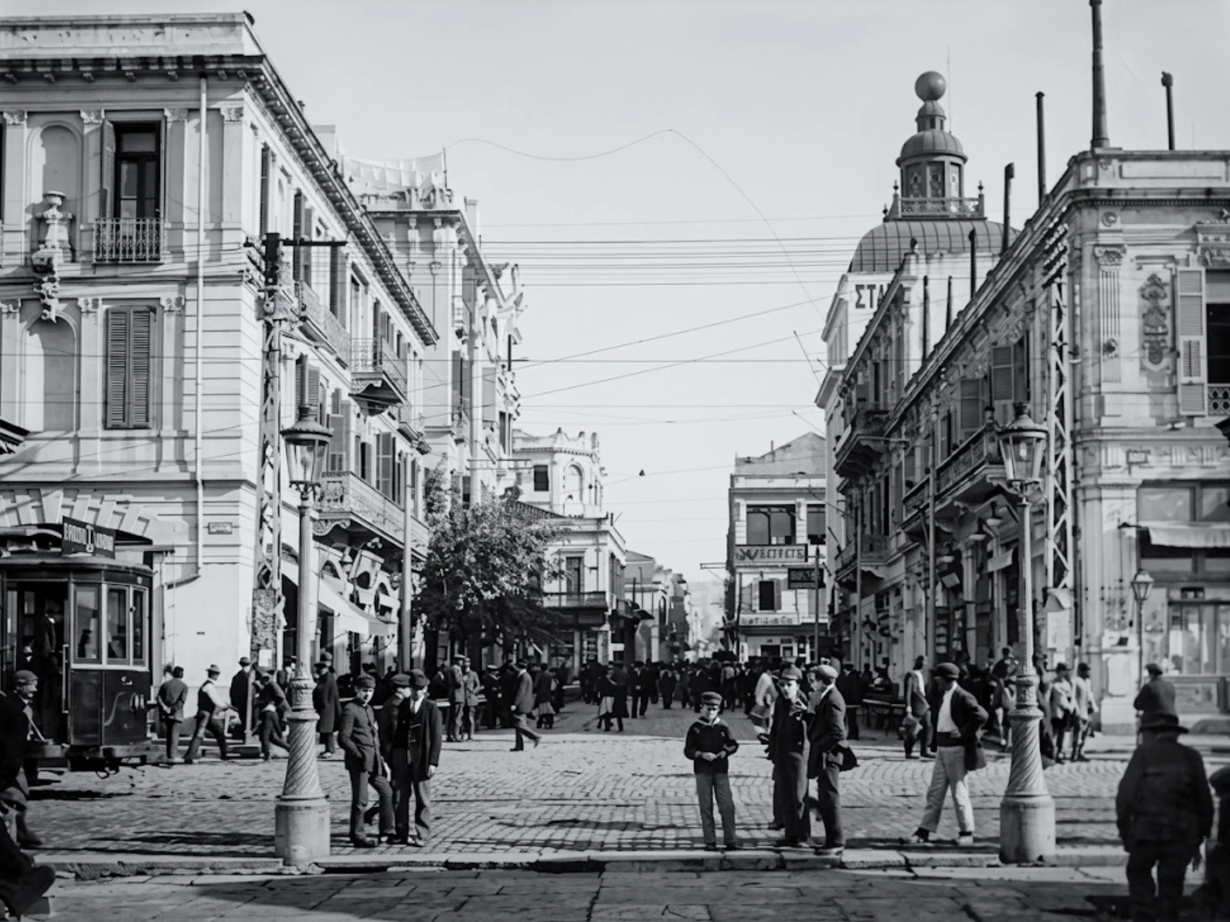 Black and white photo of a busy urban street, featuring multi-story buildings, streetcars, streetlamps, and groups of people on cobblestone paths.
